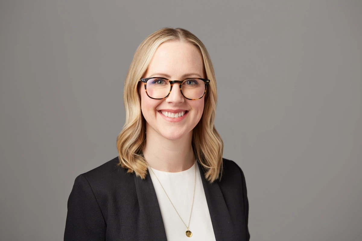 Professional headshot of female executive with natural smile, glasses, black jacket, white shirt on a gray background taken in a Vancouver office