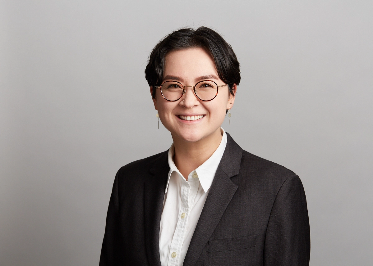 professional corporate headshot of non-binary person in dark jacket and white shirt with glasses on a grey background