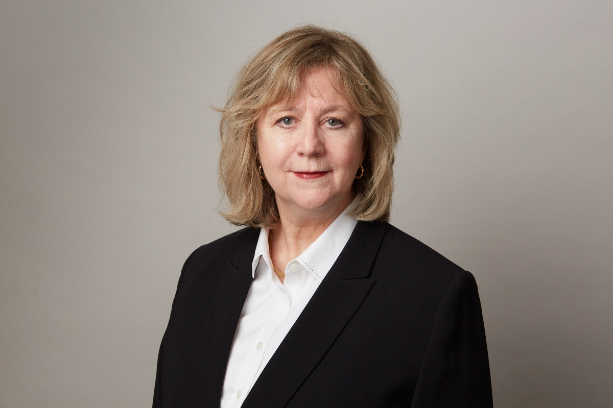 Professional corporate photograph of female board member in black jacket and white blouse on dark grey background